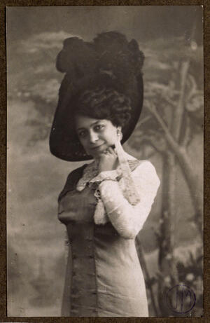 Con sombrero de ala ancha con plumas, y el rostro inclinado apoyando la barbilla en la mano